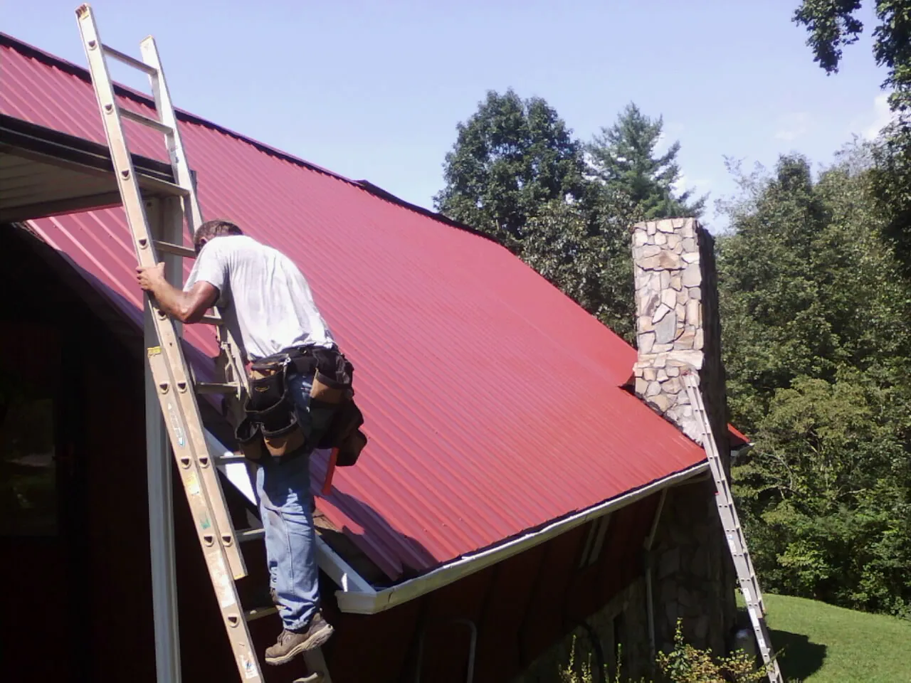 Roofer on a ladder beside a red metal roof and stone chimney — Soffit & Fascia Repair by Roofing Brothers in Cokeburg